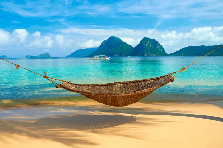 Woven hammock hanging over sandy beach with clear turquoise water and green mountains in the background under a blue sky with clouds.