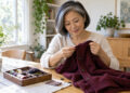 Midlife woman inspecting a burgundy cardigan beside a small sewing kit