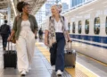 Two women pulling small suitcases along a Japanese-style railway platform.